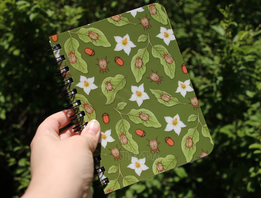 Notebook with nature-themed potato beetle cover held against a green leafy background
