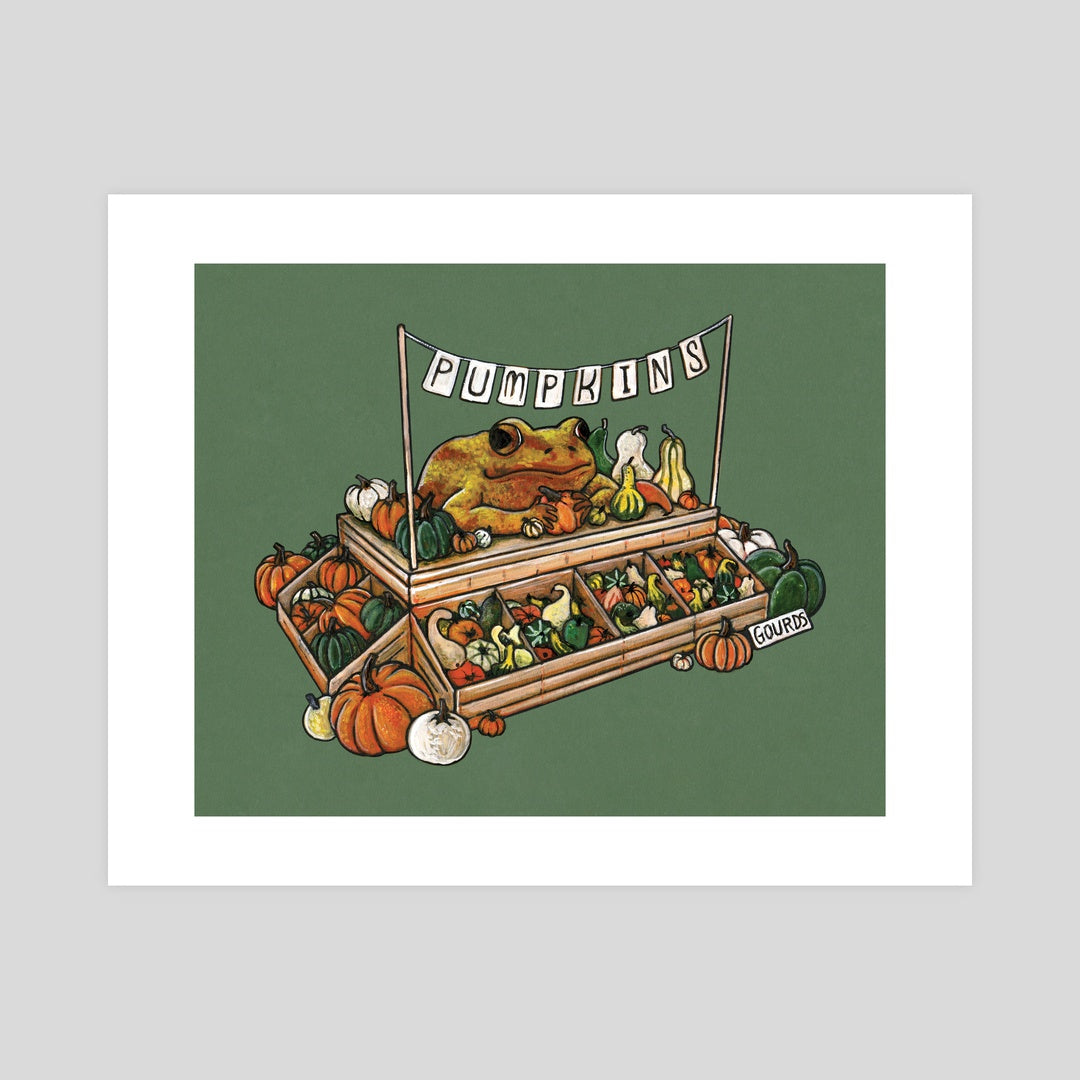 A mockup of an art print showing an illustration of a frog selling pumpkins and gourds at a market stand. The banner above the frog reads "Pumpkins".