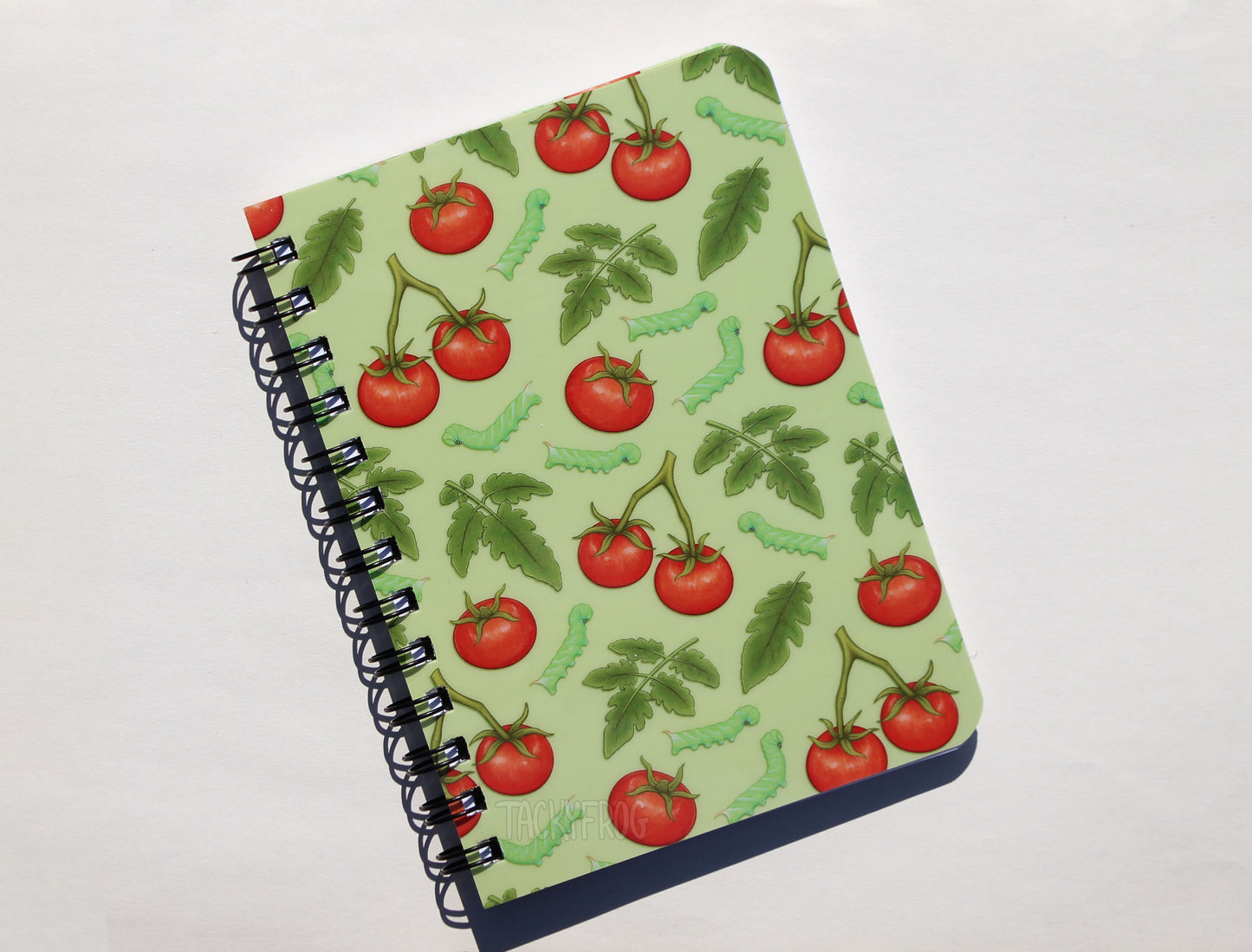 A light green 5x7 inch notebook featuring a pattern of tomatoes, tomato leaves, and tomato hornworm caterpillars.