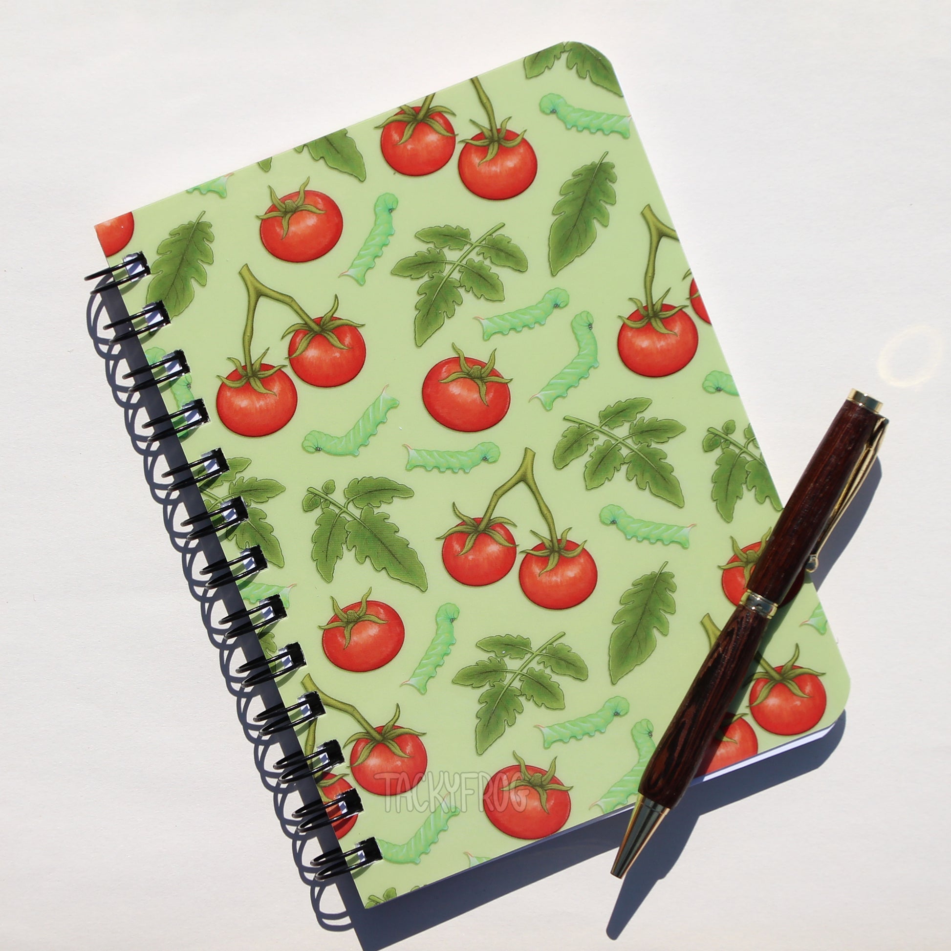 A 5 x 7 inch laminated notebook featuring a colorful pattern of tomatoes, leaves, and hornworm caterpillars on the cover. The pen is not included.