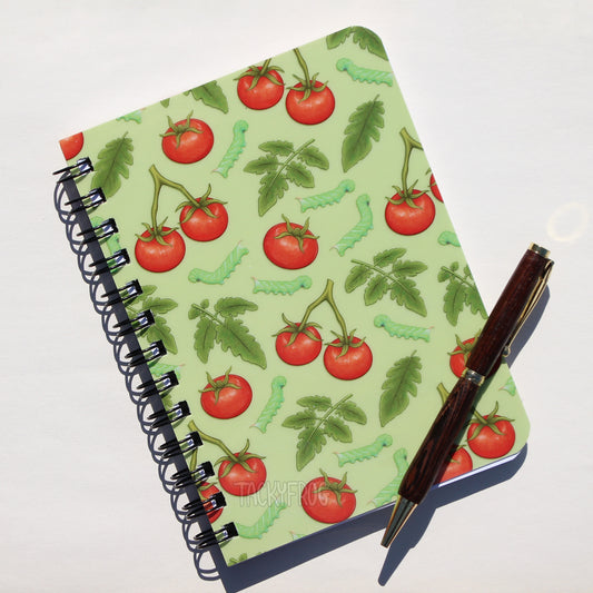 A 5 x 7 inch laminated notebook featuring a colorful pattern of tomatoes, leaves, and hornworm caterpillars on the cover. The pen is not included.