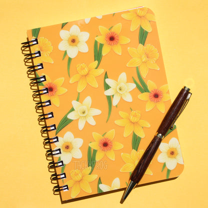 A yellow notebook featuring a pattern of yellow and white daffodils and green leaves against a yellow background. The pen is not included.