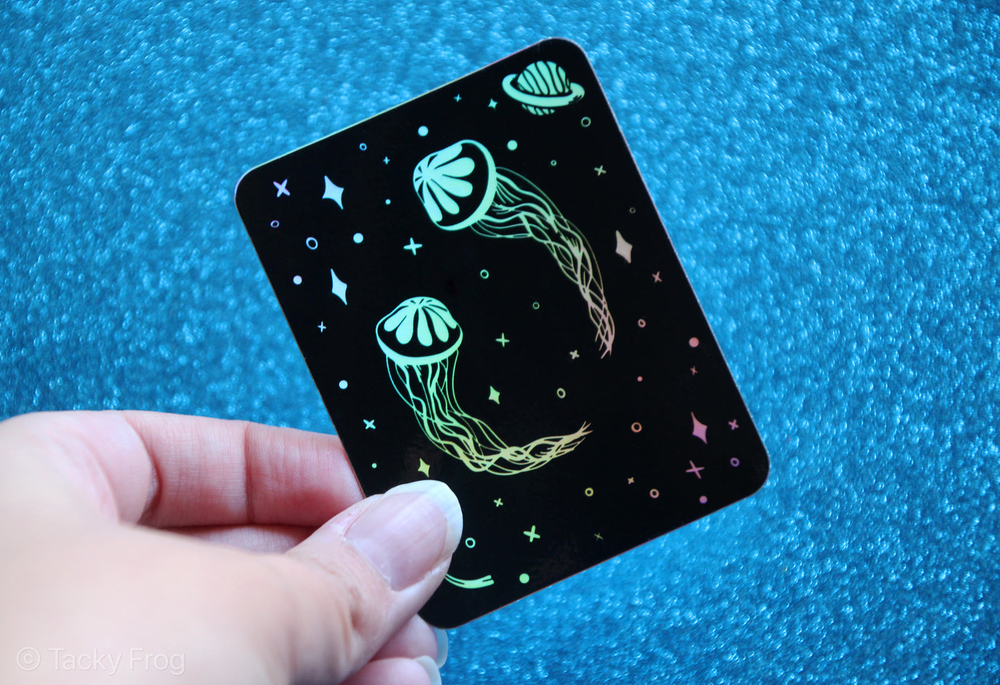 Another view of the space jellyfish holographic sticker held over blue glitter paper.