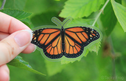 A clear monarch butterfly sticker held up in front of leaves.