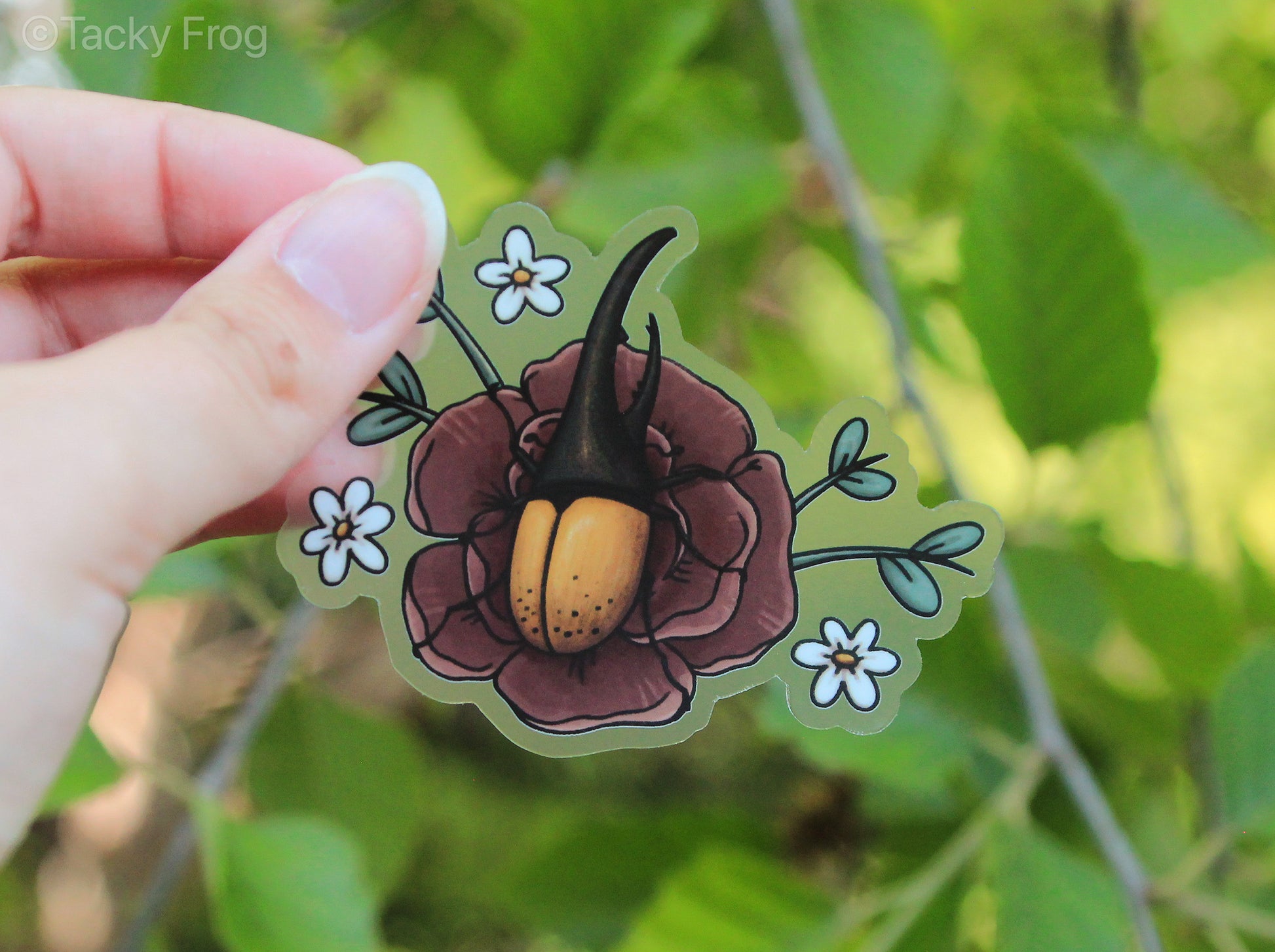 A clear sticker of a Hercules beetle on a peony flower. The peony is surrounded by small white flowers and leaves.