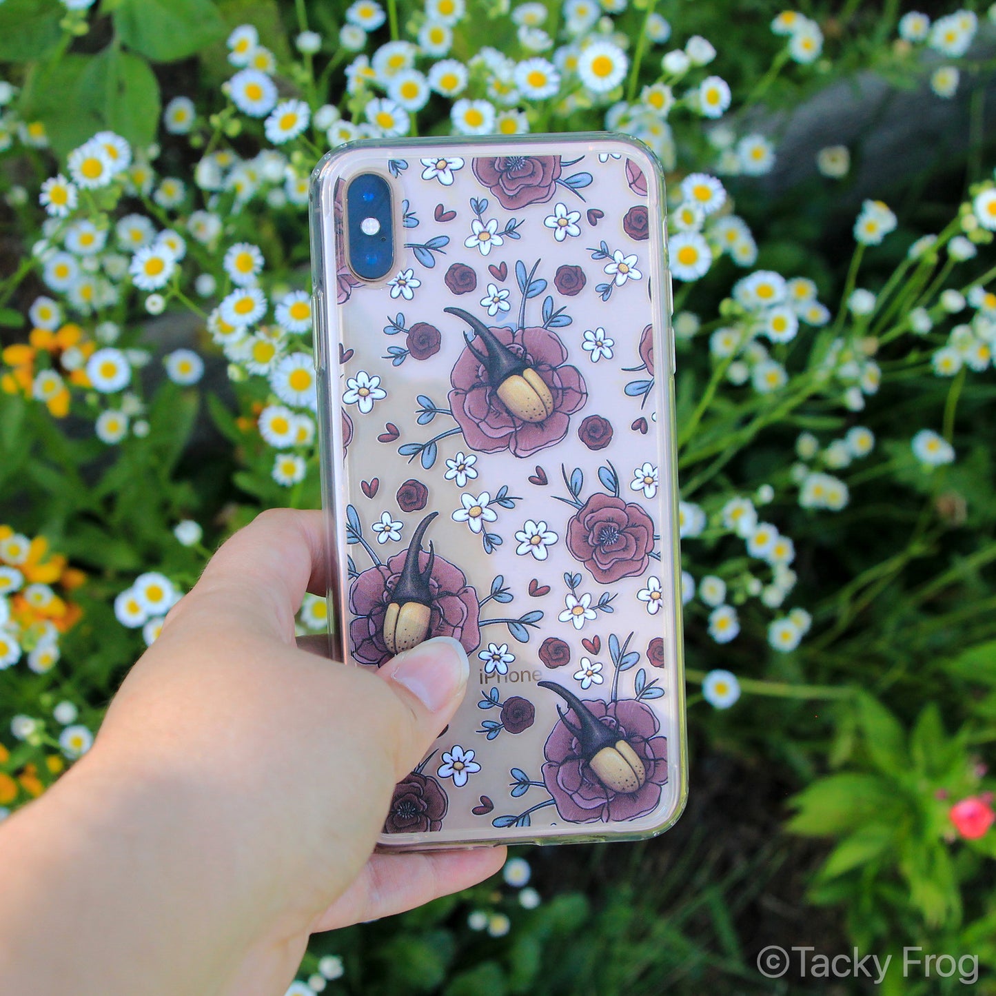 A clear Hercules Beetle and Flowers pattern phone case shown on a gold iPhone. It is held over a garden.