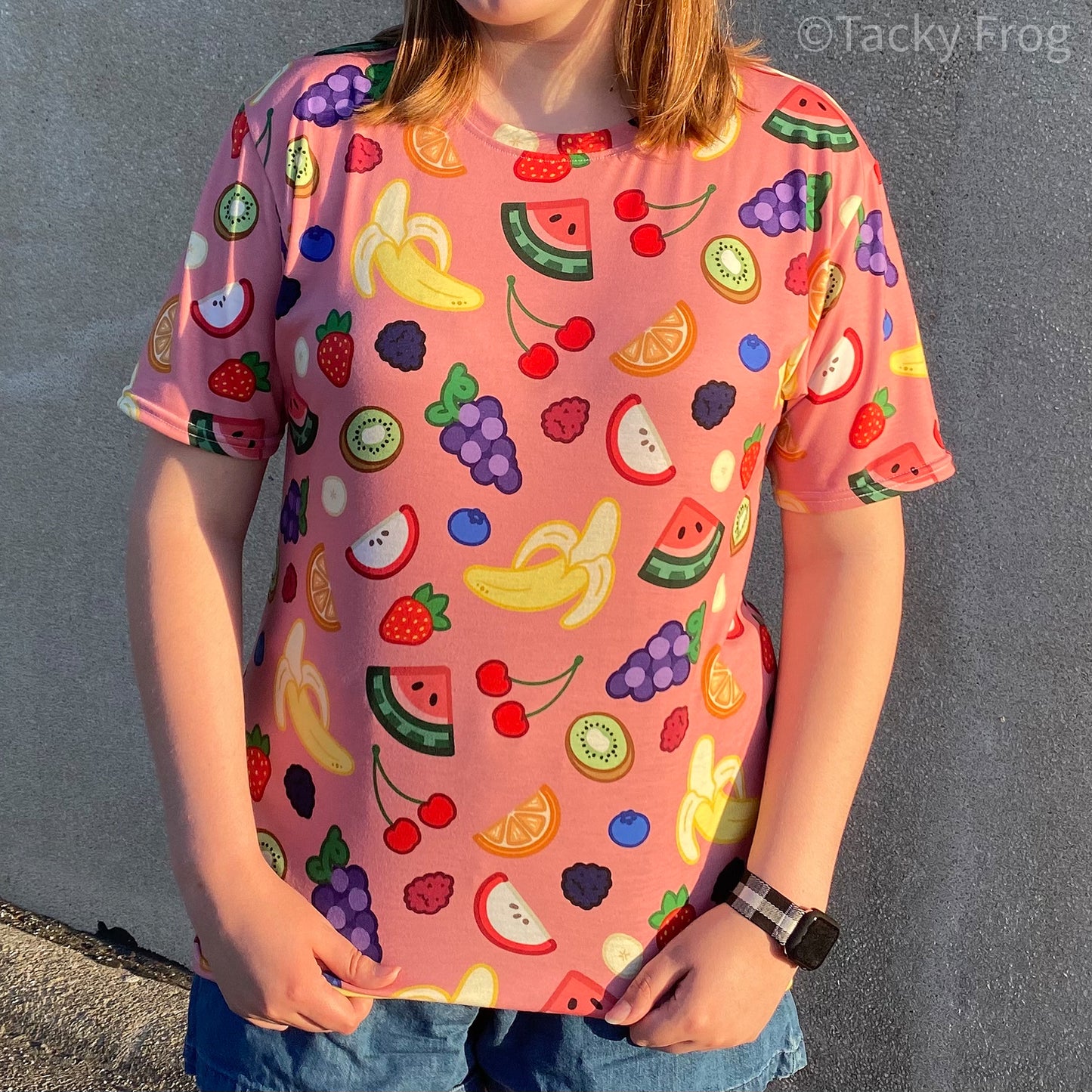 A woman wearing the fruit salad pattern t-shirt. The pattern includes illustrations of cherries, bananas, grapes, watermelon, strawberries, blueberries, oranges, blueberries, blackberries, kiwi, and apple slices. 