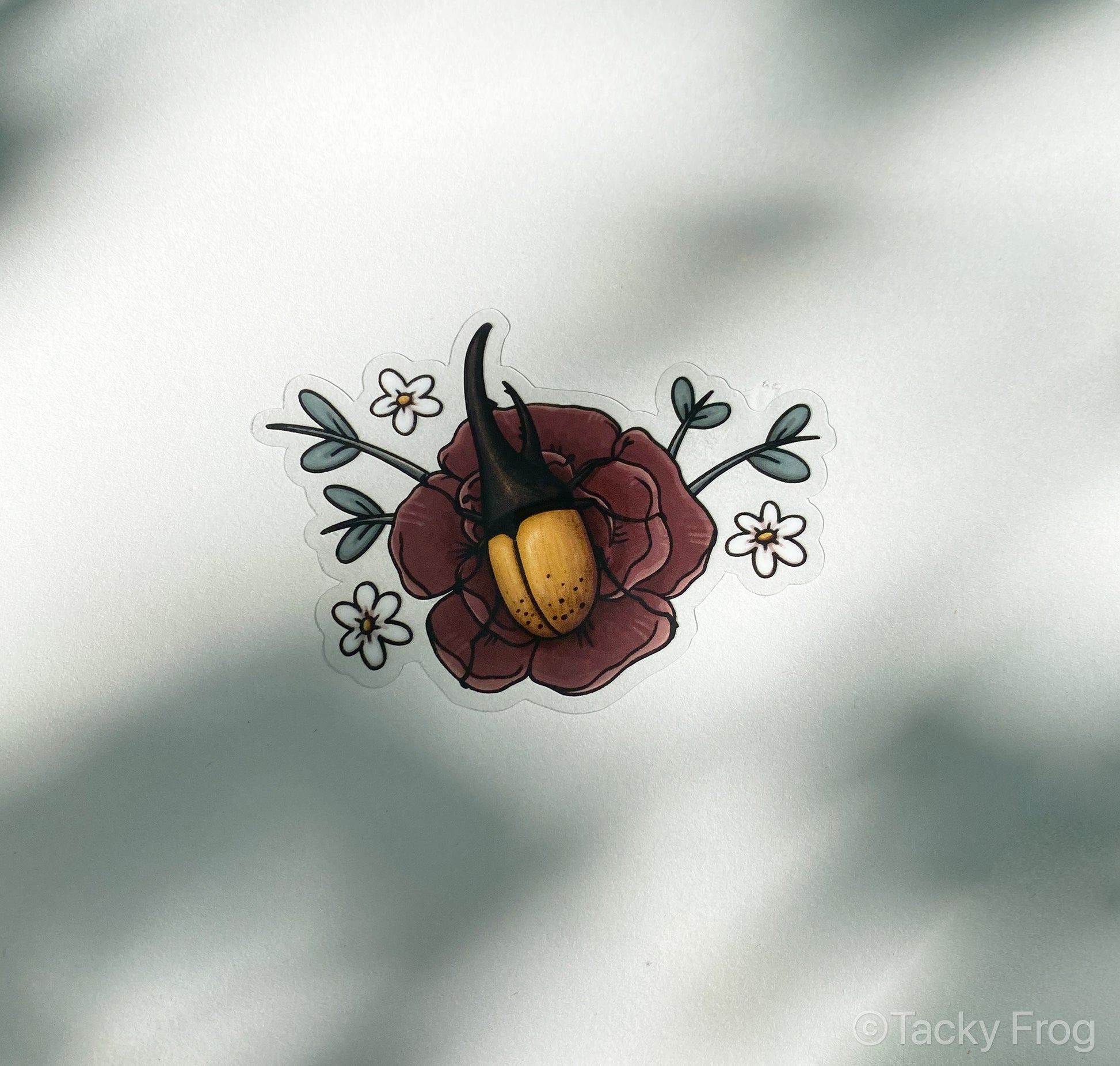 The clear Hercules beetle sticker placed on paper outdoors. The leaves are casting shadows on the paper.