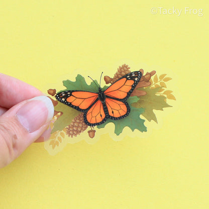 A clear vinyl sticker of a monarch butterfly perched on autumn leaves, pinecones, and acorns.