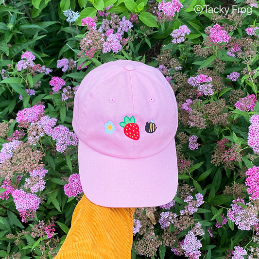 A pink hat with a strawberry, a daisy, and a bee embroidered on it.