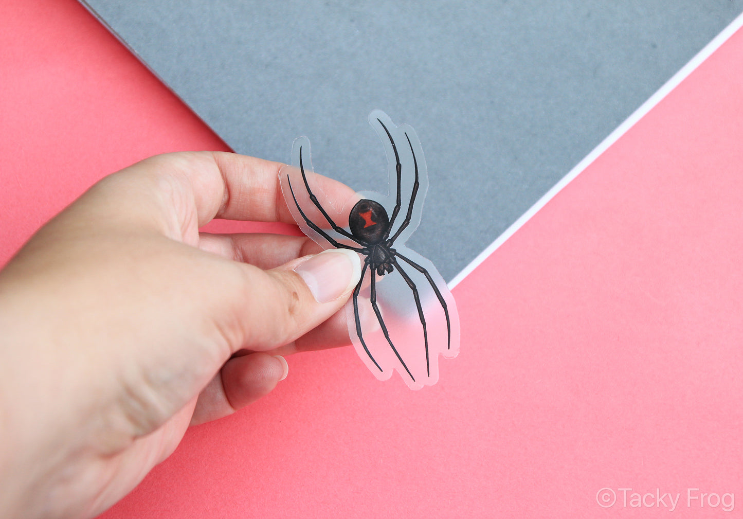 A clear vinyl sticker of a black widow spider held over a notebook.