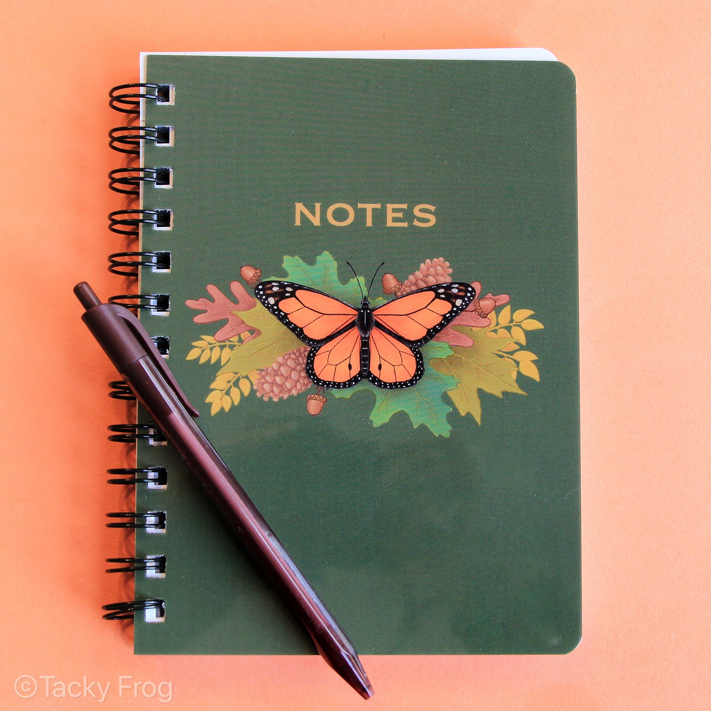 A green notebook with a glossy, laminated cover that depicts a monarch butterfly sitting on top of leaves and pinecones. The text above the design says "Notes". The pen is not included.