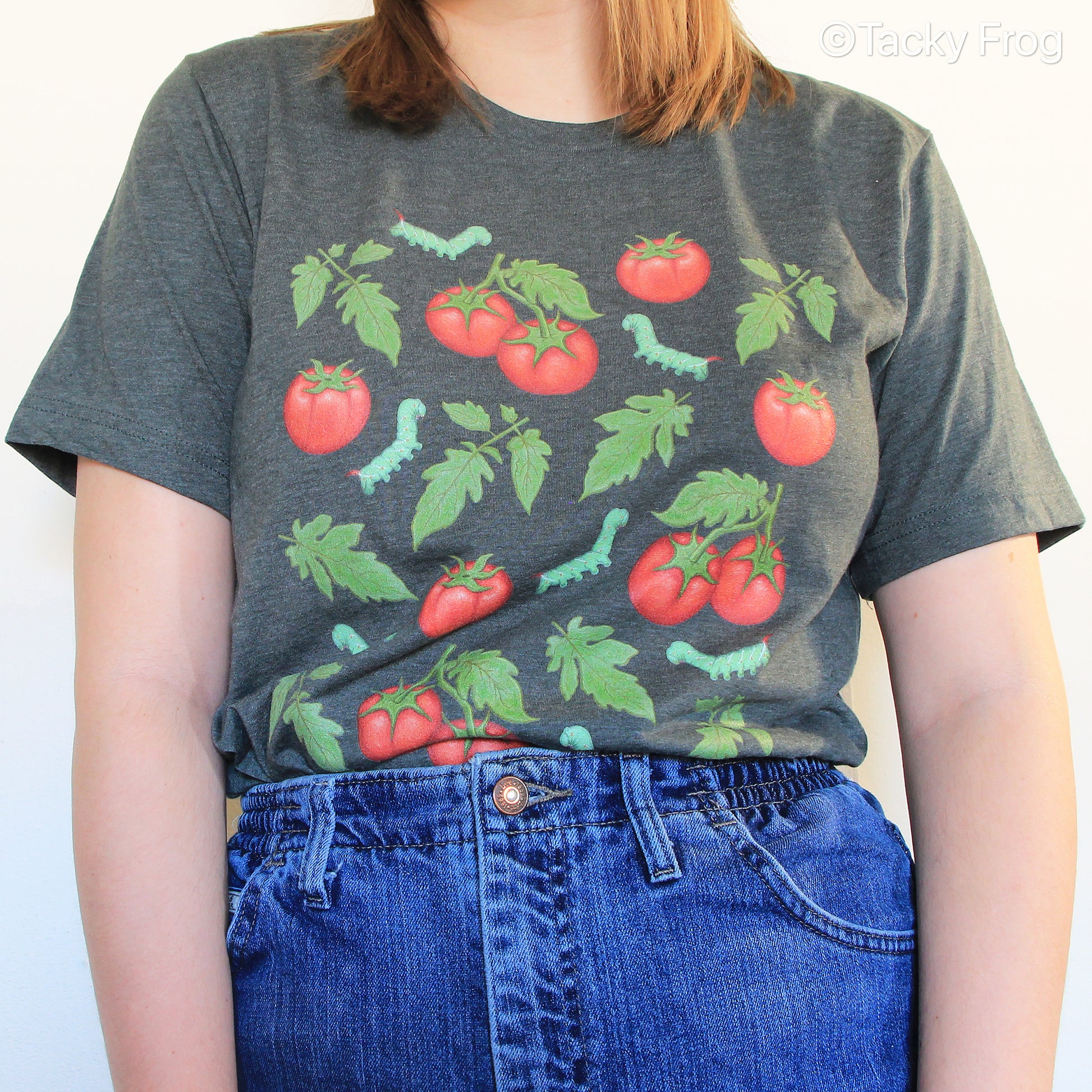 A woman wearing a heather forest-colored t-shirt with tomatoes, green caterpillars, and leaves on it. The shirt is tucked into the jeans.