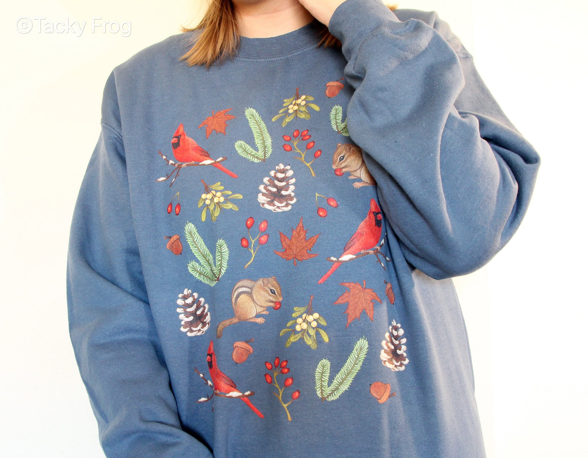 A close up of the design on the winter foraging sweatshirt. There are snowy pinecones, mistletoe, cardinals, chipmunks, rose hips, leaves, acorns, and pine tree branches.