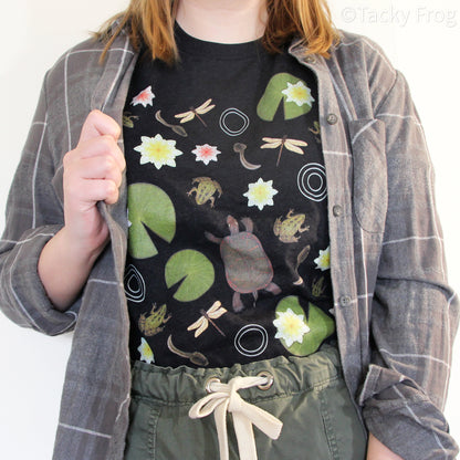 A woman wearing a very dark grey t-shirt with pond-dwelling wildlife on it such as frogs, turtles, tadpoles, and lily pads. She is wearing a grey flannel over the shirt.