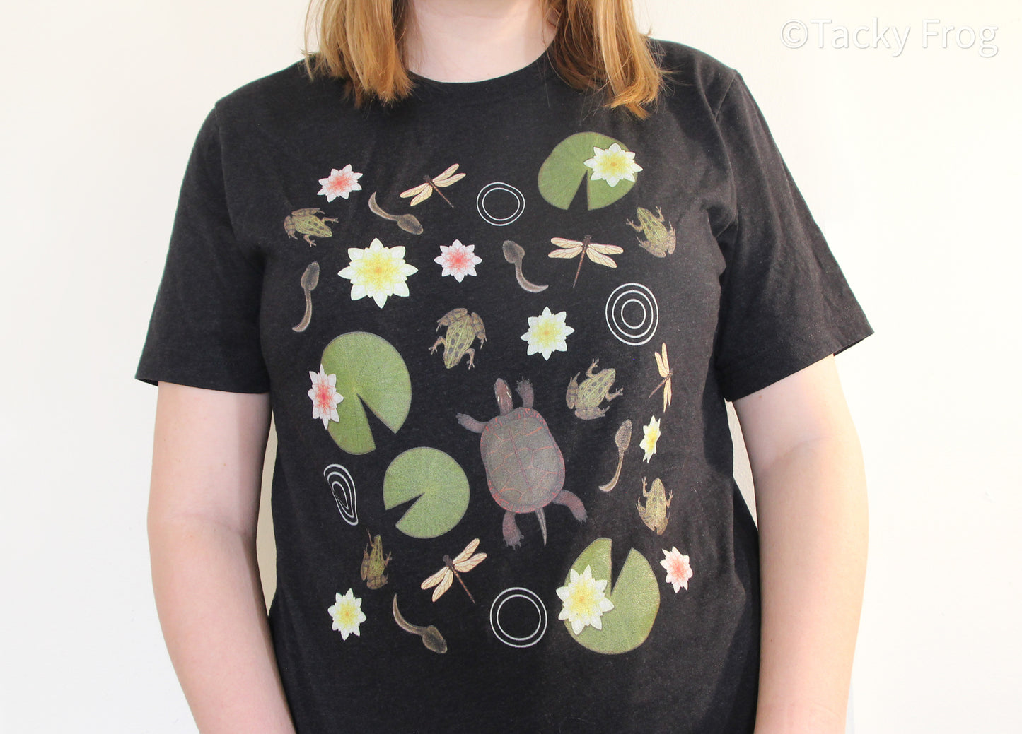 In addition to frogs, turtles, lily pads, and tadpoles, the t-shirt features lily flowers, dragonflies, and water ripples.