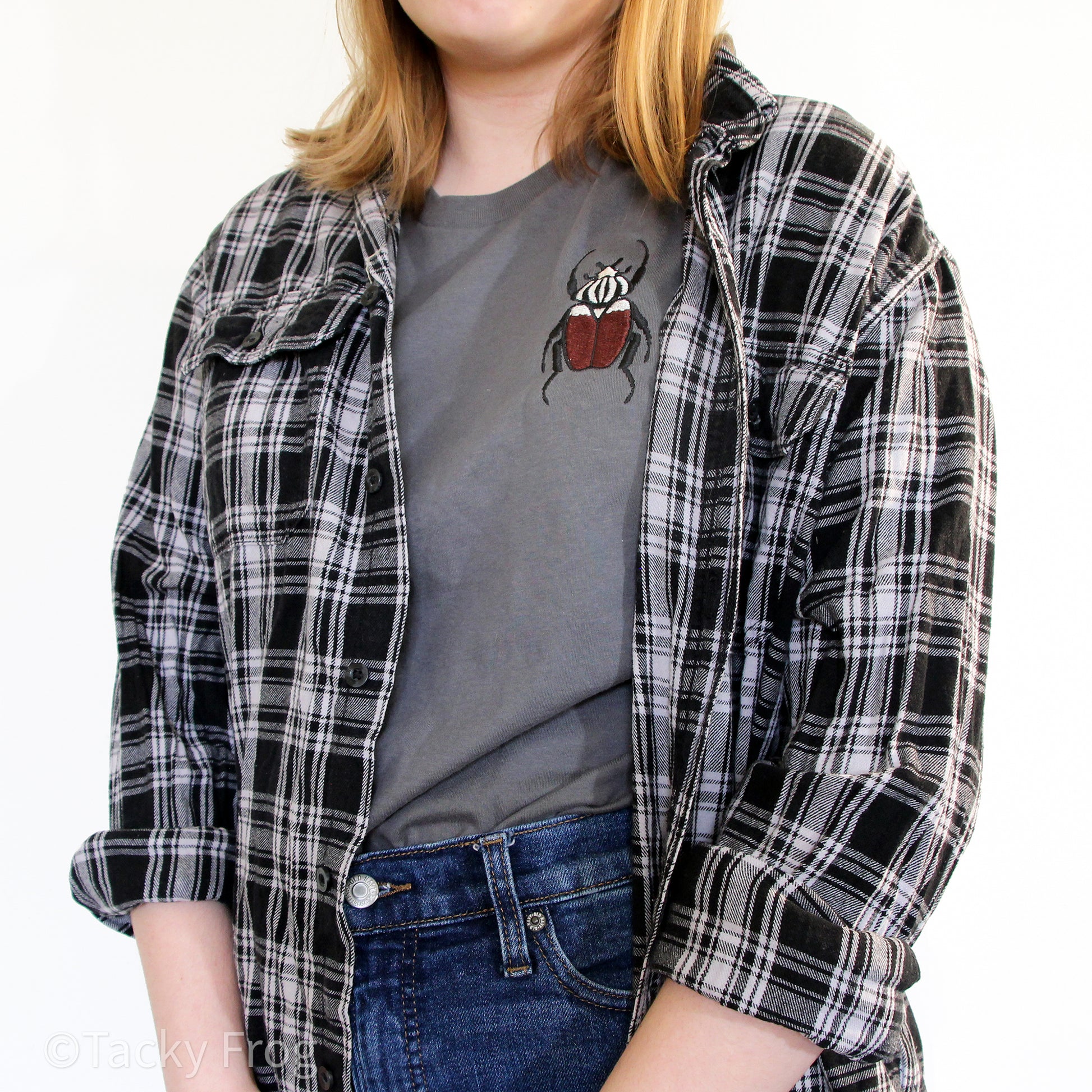 A woman wearing a t-shirt with a Goliath beetle embroidered on it. She is wearing the shirt tucked-in with a black-and-white flannel over the shirt.