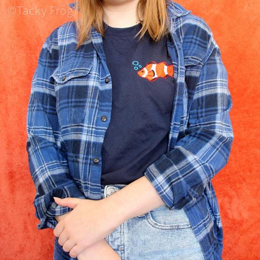 A woman wearing a navy blue t-shirt with an embroidered clownfish on it. The shirt is worn under a blue flannel.