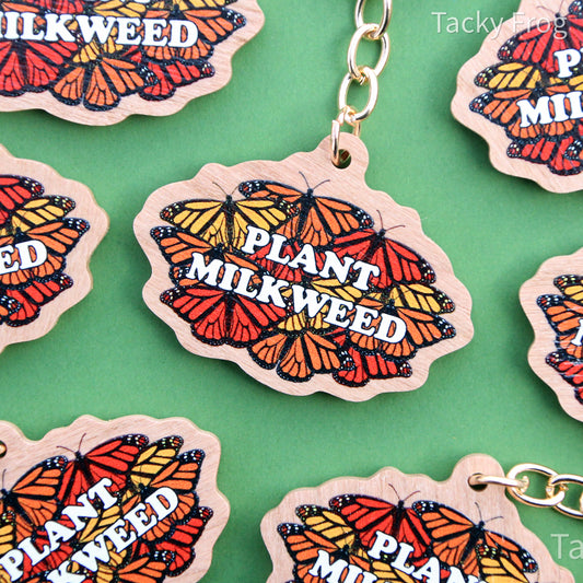 A wooden keychain depicting a design made up of monarch butterflies behind white text that reads "plant milkweed". There is a gold-colored chain attached to the charm.