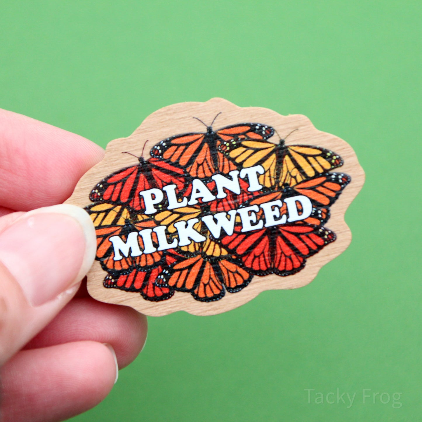 A wooden pin depicting a design made up of monarch butterflies behind white text that reads "plant milkweed".