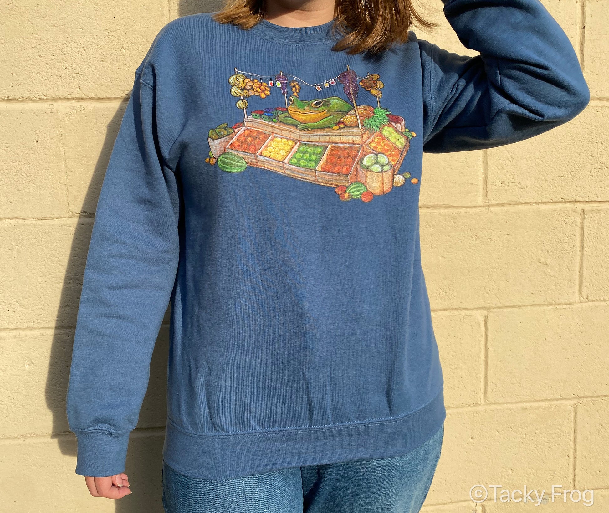 Another photo of a woman wearing an indigo-blue sweatshirt of a frog selling at a fruit stand.