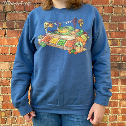 A woman wearing an indigo blue-colored sweatshirt featuring a design that depicts a frog selling fruit at a fruit stand