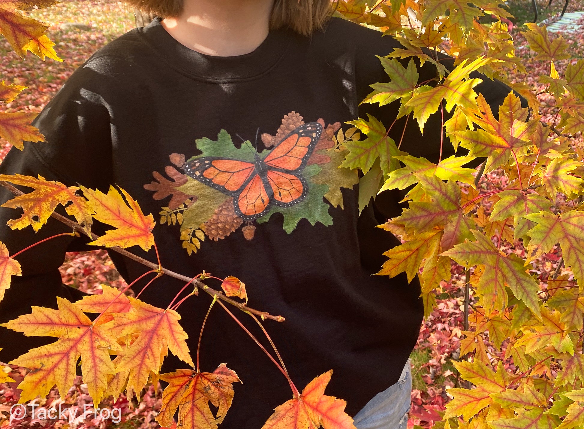 A girl wearing a black sweatshirt with a monarch butterfly on it while standing behind leaves.