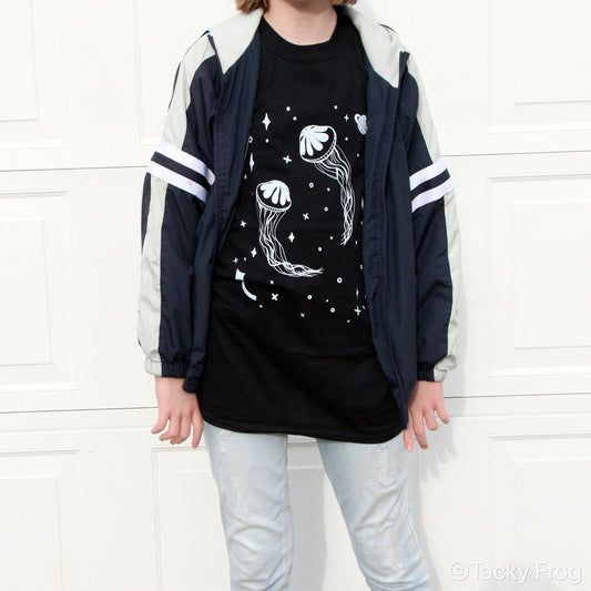 A young woman wearing a black t-shirt with jellyfish in outer space on it. She is wearing a jacket over the shirt.